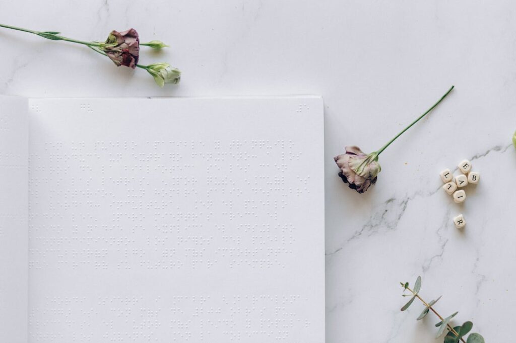 A minimalistic flatlay featuring an open braille book, dry flowers, and letter blocks on a marble surface.